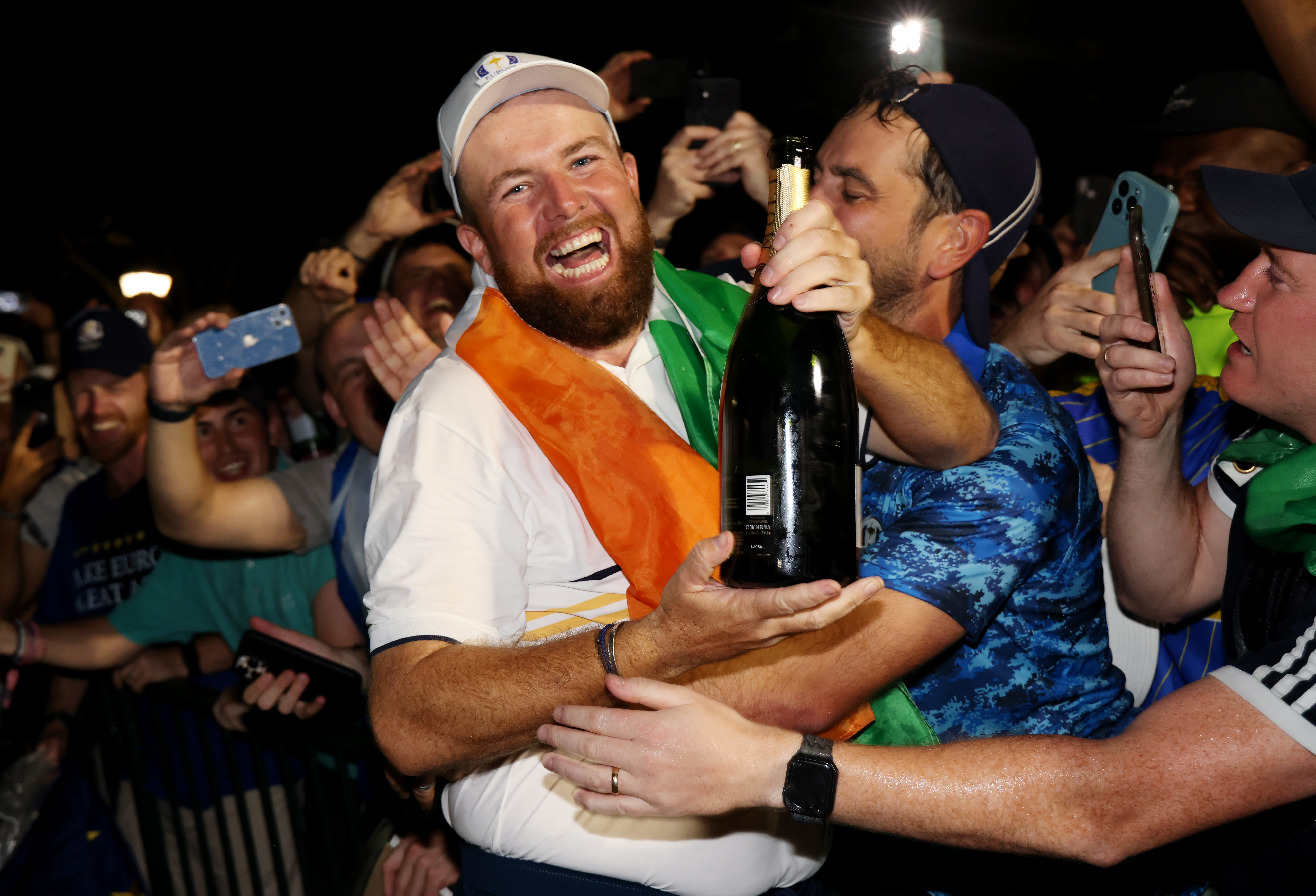 Shane Lowry smiles broadly while holding a bottle of champagne and being embraced by European fans at Bethpage